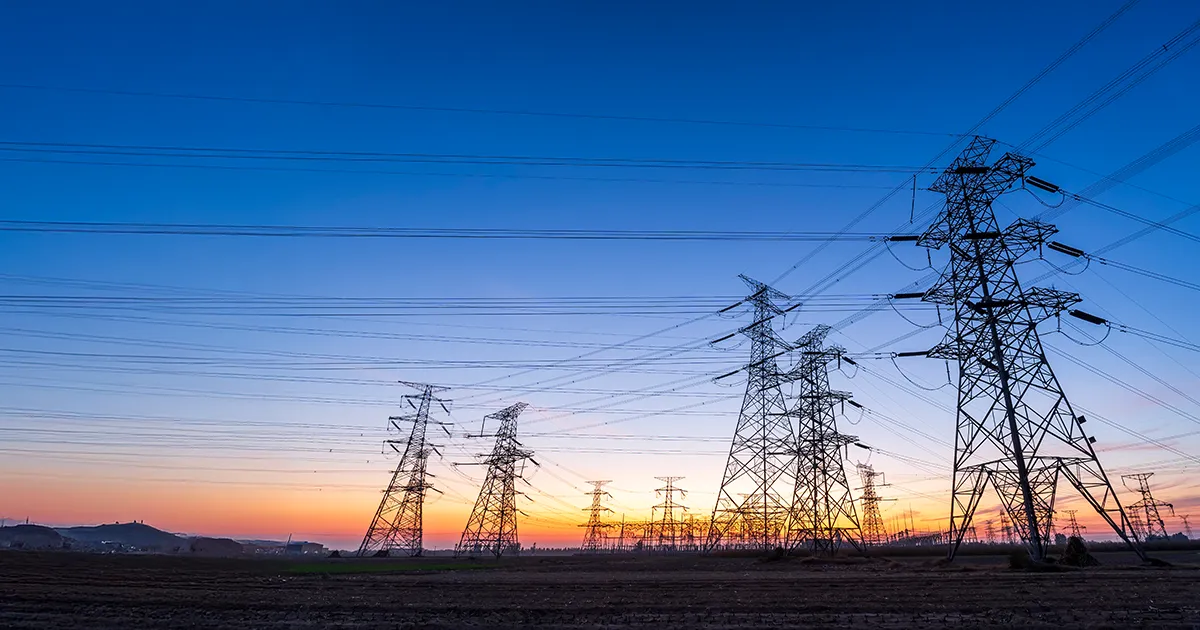Power lines from transmission towers leading into a substation are backlit by a sunset and dark blue sky.